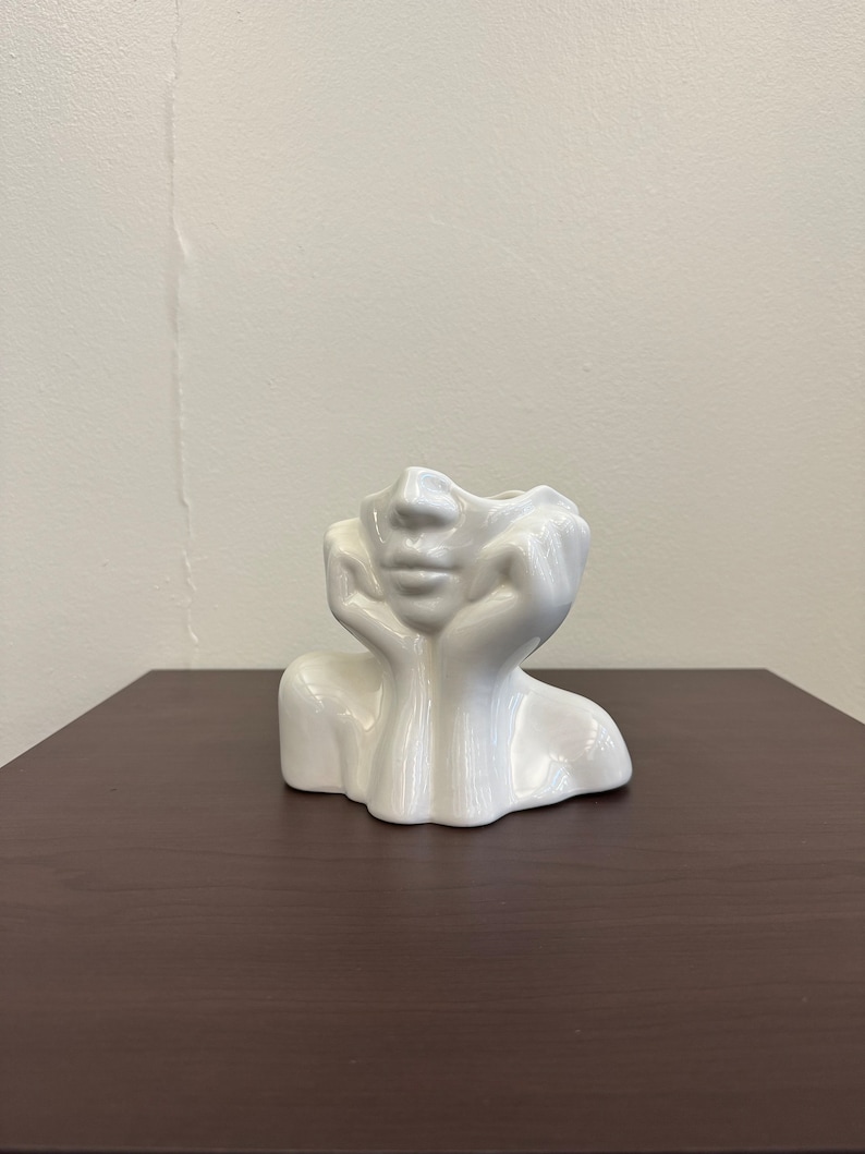 May include: A white ceramic vase shaped like a human face, with hands supporting the head. The vase has a smooth, glossy finish and sits on a dark brown surface. The face has a serene expression.
