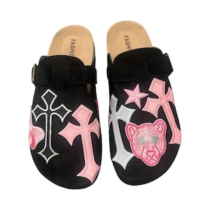May include: A pair of black suede clogs with a pink and silver cross design, a pink star, and a pink panther graphic. The clogs have a buckle strap and a cork footbed.