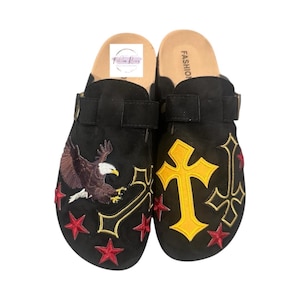 May include: A pair of black suede clogs with embroidered designs. One clog features a yellow cross and the other features a red, white, and blue eagle. Both clogs have red stars and a black strap.