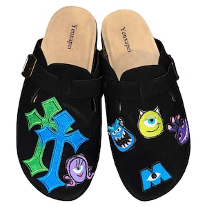 May include: A pair of black suede clogs with a buckle closure. The clogs are decorated with embroidered patches featuring characters from the movie Monsters Inc., including Mike Wazowski, Sulley, and Boo. The clogs also feature a green and blue cross design.