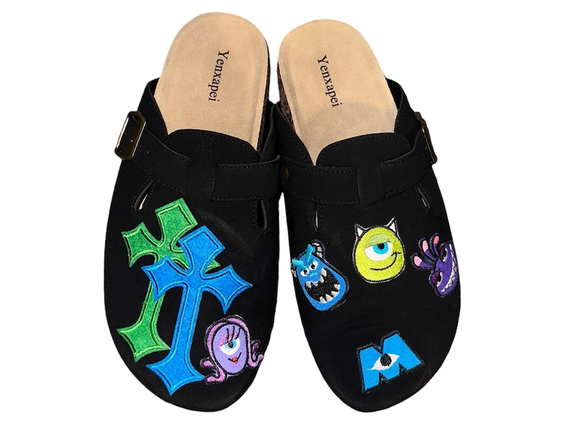 May include: A pair of black suede clogs with a buckle closure. The clogs are decorated with embroidered patches featuring characters from the movie Monsters Inc., including Mike Wazowski, Sulley, and Boo. The clogs also feature a green and blue cross design.