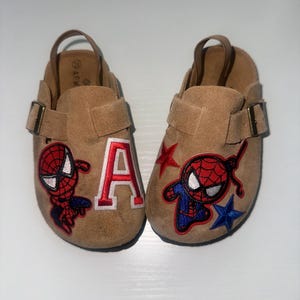 May include: A pair of tan suede clogs featuring adjustable straps with bronze-toned buckles and heel straps. The shoes are decorated with embroidered patches of a red and blue superhero character, a white and red letter A, and red and blue stars.