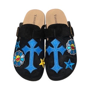 May include: A pair of black suede clogs with a blue cross, yellow star, and blue flower embroidery. The clogs have a buckle strap and the words "Yenxap" are printed on the insole.