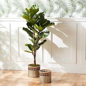 May include: A large green artificial fiddle leaf fig tree in a brown metal planter with a decorative metal grid pattern. The plant is sitting on a light wood floor in front of a white wall with a green tropical leaf wallpaper.