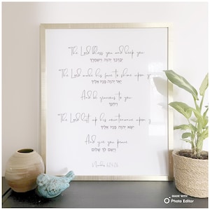 May include: A white framed print with a gold border featuring a religious quote in English and Hebrew. The quote is from Numbers 6:24-26. The print is on a white background and has a simple, elegant design.