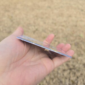 May include: A rectangular card with a glossy finish, held in a hand. The card has a colorful design on the front, with visible edges. The background is blurred, suggesting an outdoor setting.
