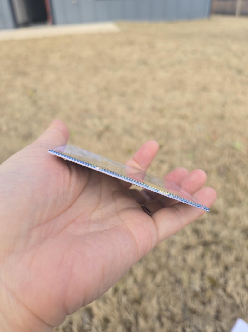 May include: A rectangular card with a glossy finish, held in a hand. The card has a colorful design on the front, with visible edges. The background is blurred, suggesting an outdoor setting.