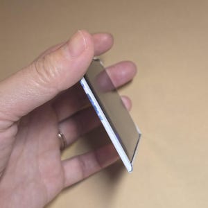 May include: A rectangular magnet with a white border and a glossy black surface. The edge of the magnet shows a hint of blue and white design. The magnet is held in a hand.