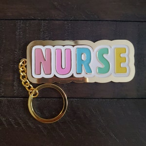 以下が含まれることがあります： パステルカラーで「NURSE」の文字が入ったゴールドカラーのキーホルダー。文字はピンク、ブルー、グリーン、イエローで、白とゴールドで縁取られています。キーホルダーにはゴールドのチェーンと丸いキーリングが付いています。