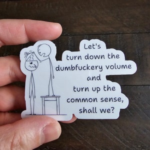 May include: A white sticker with a cartoon illustration of two stick figures. One figure stands on a small table, adjusting the other's head. Text reads: "Let's turn down the dumbfuckery volume and turn up the common sense, shall we?"