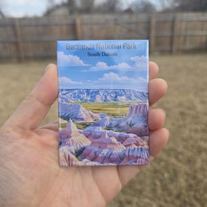 Puede incluir: Un imán rectangular con un paisaje del Parque Nacional Badlands en Dakota del Sur. La imagen muestra una colorida vista de formaciones rocosas en capas bajo un cielo azul con nubes. El texto "Badlands National Park South Dakota" está impreso en la parte superior.