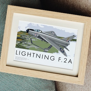 May include: A framed print of a vintage illustration of a silver and gray English Electric Lightning F.2A supersonic jet fighter plane. The plane is flying over a green field with a runway and buildings in the background. The print has the text "LIGHTNING F.2A" in black letters.