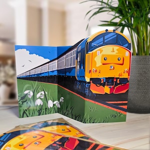 May include: A pop-up card depicting a yellow and blue train on a green field with white flowers. The card is open, revealing a 3D illustration of the train against a blue sky. The card is designed to stand upright.