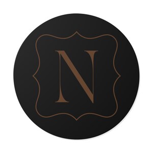 Könnte beinhalten: Ein runder, schwarzer Untersetzer mit einem braunen Zierrand und dem braunen Buchstaben "N" in der Mitte. Der Buchstabe ist in einer Serifenschrift gehalten. Der Untersetzer hat ein schlichtes, elegantes Design.