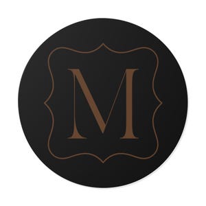 Könnte beinhalten: Ein runder, schwarzer Untersetzer mit einem braunen Zierrahmen und dem braunen Buchstaben "M" in der Mitte. Das Design ist schlicht und elegant, passend für verschiedene Einrichtungsstile.