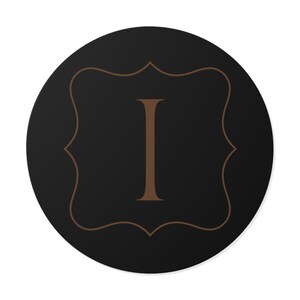 Op de afbeelding: Een ronde, zwarte decoratieve sticker met een bruine letter "I" in een decoratief bruin kader. Het kader heeft een geschulpte rand. De sticker staat op een witte achtergrond.