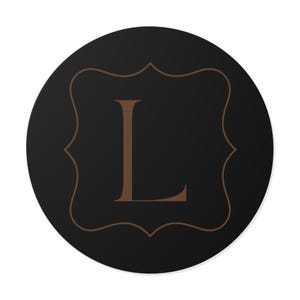 Könnte beinhalten: Ein runder, schwarzer Untersetzer mit einem braunen, verzierten Rahmen und dem Buchstaben "L" in der Mitte. Der Buchstabe und der Rahmen haben ein klassisches, elegantes Design. Der Untersetzer ist wahrscheinlich aus einem haltbaren Material.