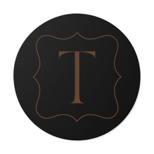 Könnte beinhalten: Ein runder, schwarzer Untersetzer mit einem braunen, verzierten Rahmen und dem Großbuchstaben "T" in der Mitte. Das Design ist schlicht und elegant, passend für verschiedene Einrichtungsstile.