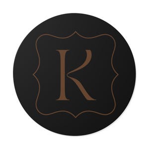 Könnte beinhalten: Ein runder, schwarzer Untersetzer mit einem braunen, verzierten Rahmen und dem Buchstaben "K" in der Mitte. Der Buchstabe und der Rahmen sind in einem warmen Braunton gehalten, der sich vom schwarzen Hintergrund abhebt. Der Untersetzer hat ein schlichtes, elegantes Design.