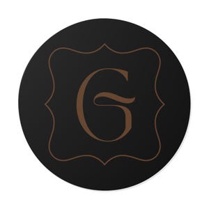 May include: A round, black coaster with a brown decorative border and a brown letter "G" in the center. The letter is in a stylized, elegant font. The coaster has a white background.