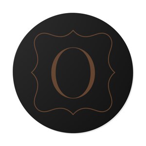 Könnte beinhalten: Ein schwarzer runder Untersetzer mit einem braunen dekorativen Rahmen und dem braunen Buchstaben "O" in der Mitte. Der Untersetzer hat ein schlichtes, elegantes Design.