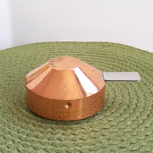 May include: A copper and silver measuring cup with a handle. The cup is round and has a conical shape. The handle is attached to the side of the cup.