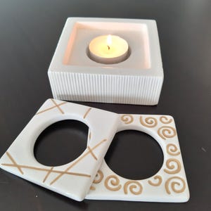 May include: White ceramic candle holder with a square base and a round opening for a tea light candle. The holder has a textured surface and is decorated with gold lines. There are two additional white ceramic candle holders with gold designs, one with a criss-cross pattern and the other with a swirl pattern.