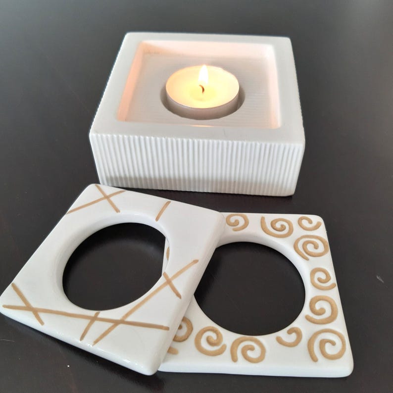 May include: White ceramic candle holder with a square base and a round opening for a tea light candle. The holder has a textured surface and is decorated with gold lines. There are two additional white ceramic candle holders with gold designs, one with a criss-cross pattern and the other with a swirl pattern.