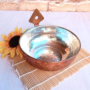 May include: A hammered copper bowl with a handle. The bowl is shiny and has a light blue tint. It is sitting on a bamboo mat.