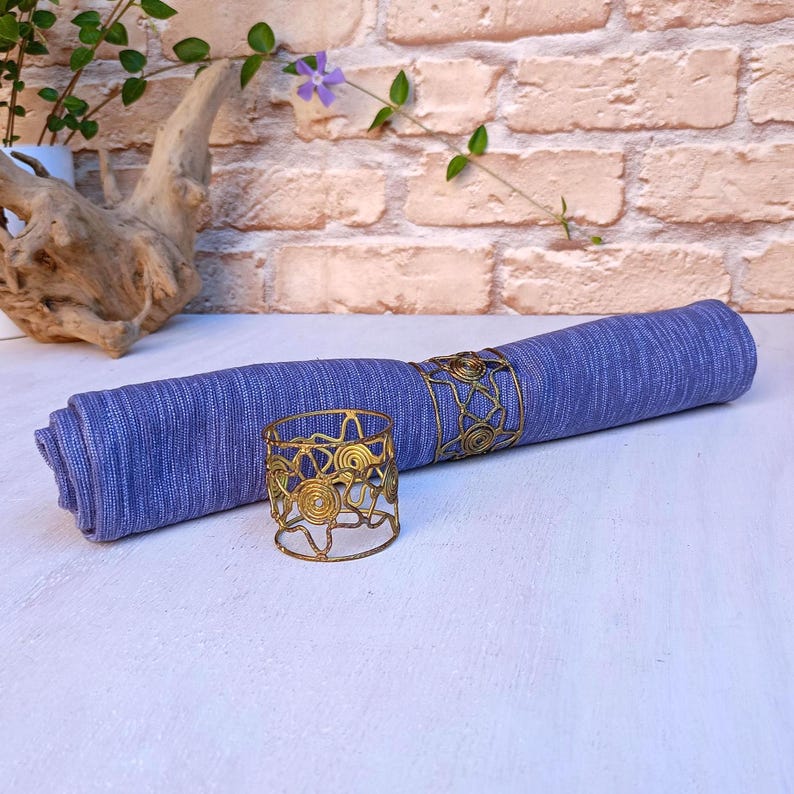 Pu&ograve; includere: Una tovagliolo di lino blu arrotolato con un anello portatovagliolo in metallo color oro. Un altro anello portatovagliolo dorato &egrave; accanto al tovagliolo arrotolato. Gli anelli portatovaglioli hanno un design traforato. Lo sfondo include un muro di mattoni e verde.