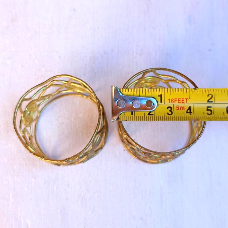 Pu&ograve; includere: Due anelli portatovaglioli in metallo traforato color oro. Un anello &egrave; misurato con un metro a nastro, che mostra un diametro di circa 5 cm. Gli anelli hanno un design decorativo a spirale e sono su uno sfondo bianco.