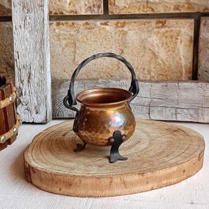 May include: A small, round, hammered copper cauldron featuring a dark, twisted metal handle and three decorative, curved black metal feet. The metallic surface displays a warm, reflective finish with visible indentations from the hammering process.