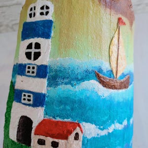 May include: Hand-painted wooden ornament featuring a lighthouse, a small house, a sailboat, and a blue ocean scene. The lighthouse is white with blue stripes and a black door. The house has a red roof and white walls. The sailboat is brown with a yellow sail and a red flag.