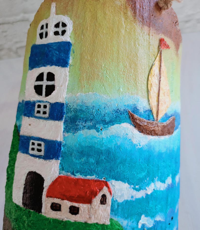 May include: Hand-painted wooden ornament featuring a lighthouse, a small house, a sailboat, and a blue ocean scene. The lighthouse is white with blue stripes and a black door. The house has a red roof and white walls. The sailboat is brown with a yellow sail and a red flag.