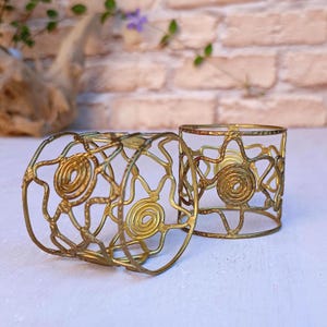 Pu&ograve; includere: Due portacandele in metallo color oro. I supporti hanno un design traforato con motivi a spirale e lineari. Un supporto &egrave; sdraiato su un lato, mentre l'altro &egrave; in posizione verticale. Lo sfondo presenta un muro di mattoni e vegetazione.