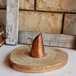 May include: A small, conical copper vessel featuring a hammered texture and a single curved handle. The object has a wide, flat circular base and an angled top opening, showcasing a polished metallic finish with warm, reflective orange-brown tones.