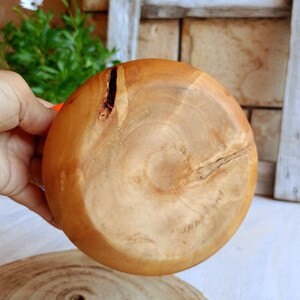 May include: A round wooden bowl with a smooth, light brown finish, featuring visible natural wood grain patterns and a small, dark, irregular knot or fissure near the upper left edge, showcasing the organic texture of the material.