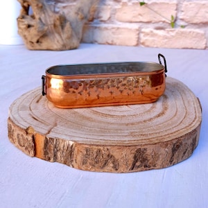 May include: A copper-coloured, hammered metal planter with black handles. The rectangular container is approximately 25 cm long and sits on a round, natural wood slice. The planter has a shiny, reflective surface.