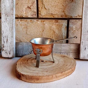 Peut inclure: Une petite casserole en cuivre avec un intérieur métallique, dotée de trois pieds en métal robustes et d'un long manche fin en métal sur le côté. L'extérieur de la casserole présente une finition en cuivre poli, avec des pieds fixés par rivets.