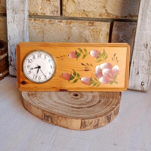 May include: A rectangular wooden clock with a light brown finish. The clock face is round with black numbers and hands. The wooden frame is decorated with pink and white floral designs. The clock sits on a wooden slice.