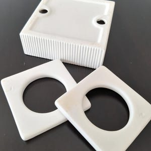 May include: Three white ceramic ring holders. The holders are square with a round hole in the center. The holders are on a black surface.