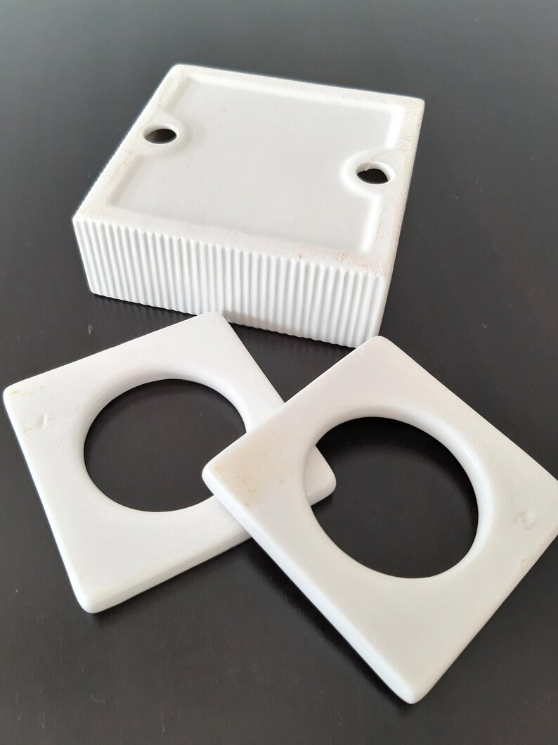 May include: Three white ceramic ring holders. The holders are square with a round hole in the center. The holders are on a black surface.