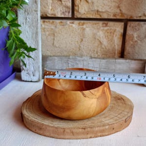 May include: A small, round, light brown wooden bowl with visible natural grain patterns. A white measuring tape is placed across the top, showing a diameter of approximately 3.5 inches. The bowl sits on a circular, light-colored wood slice base.