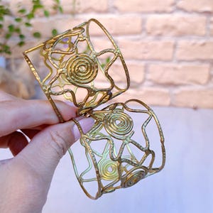 Pu&ograve; includere: Due braccialetti a polsino in metallo color oro. Ogni braccialetto presenta un design traforato con motivi a spirale e a forma di stella. I braccialetti sono tenuti su uno sfondo di muro di mattoni.