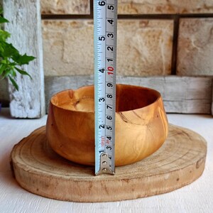 May include: A light brown wooden bowl with a smooth, rounded shape and visible natural wood grain patterns. A vertical measuring tape is placed against the bowl, indicating a height of approximately 3.5 inches from the base to the rim.