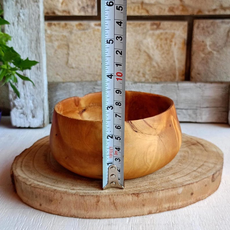 May include: A light brown wooden bowl with a smooth, rounded shape and visible natural wood grain patterns. A vertical measuring tape is placed against the bowl, indicating a height of approximately 3.5 inches from the base to the rim.