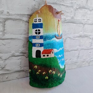 May include: A hand-painted wooden plaque depicting a lighthouse with blue and white stripes, a small house with a red roof, and a sailboat on a blue and white ocean background. The plaque has a rustic, natural wood texture.