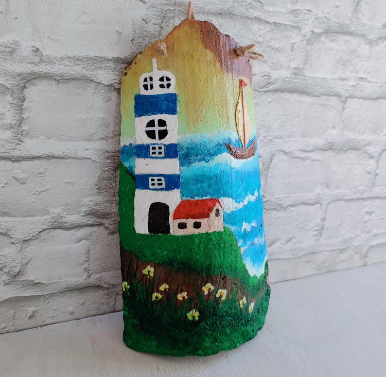 May include: A hand-painted wooden plaque depicting a lighthouse with blue and white stripes, a small house with a red roof, and a sailboat on a blue and white ocean background. The plaque has a rustic, natural wood texture.