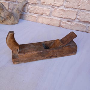 May include: A vintage wooden hand plane, featuring a curved handle and a rectangular body. The plane has a weathered, aged appearance, with visible wood grain and a metal blade. This woodworking tool is designed for smoothing and shaping wood surfaces.