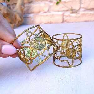 Pu&ograve; includere: Due anelli portatovaglioli in metallo dorato traforato. Uno &egrave; quadrato, l'altro cilindrico. Gli anelli presentano un motivo geometrico a spirale con spirali e forme a stella. Sono esposti su una superficie bianca.